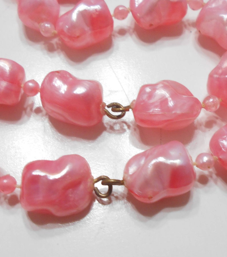 May include: A pink plastic pearl necklace with a gold clasp. The pearls are irregular in shape and have a slight iridescent sheen.