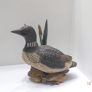May include: A hand-painted wooden sculpture of a loon bird. The bird has a black head and back with white spots, a white and black striped neck, and a tan body. It is perched on a wooden base with cattails and a green leaf behind it.