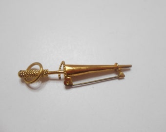 May include: A gold-toned brooch in the shape of a stylized sword with a decorative handle. The brooch has a pin on the back.