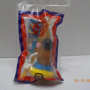 May include: A plastic Mr. Potato Head toy in a clear plastic bag. The toy is wearing a blue shirt and a black hat. The toy is holding a white plastic fishing rod. The bag has a red, white, and blue border. The text "Mr. Potato Head" is printed on the bag.