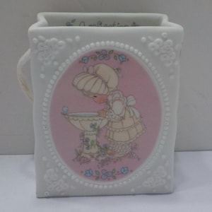 May include: White ceramic mug with a rectangular shape and a handle. The mug features a central oval image of a child in a dress looking at a birdbath. The image is framed by a pink background and a white border. Floral details adorn the mug.