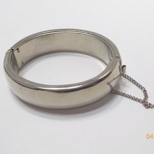 May include: A silver-toned hinged bangle bracelet. The bracelet has a rounded shape with a clasp and a safety chain. The surface is smooth and reflective. The bracelet is a piece of jewelry.