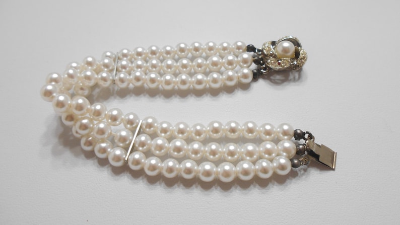 May include: A triple strand pearl bracelet with a decorative clasp featuring a faux pearl and rhinestones.