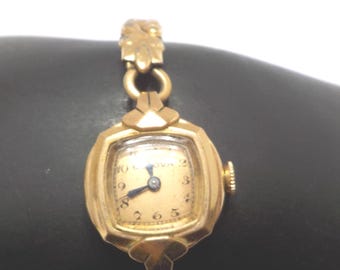 May include: A vintage gold-toned Bulova wristwatch with a square face and a delicate linked band. The watch features a cream-colored dial with black numerals and hands. The watch is an elegant accessory.
