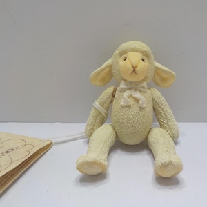 May include: A cream-colored lamb figurine with a bow tie. The lamb has large ears and is sitting with its legs extended. A tag is attached to the lamb with a string. The lamb is a decorative item.