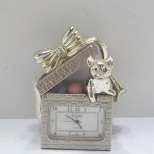May include: Silver-toned cat-themed quartz clock in the shape of a gift box. The clock face reads "Elgin" and displays the time. A decorative bow sits atop the box, and a small cat figure is perched on the edge. Inside the box are small balls of yarn.