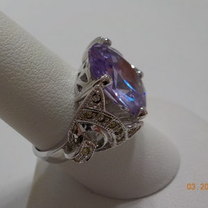 May include: A silver ring with a large, oval-shaped, purple gemstone surrounded by smaller, clear gemstones.