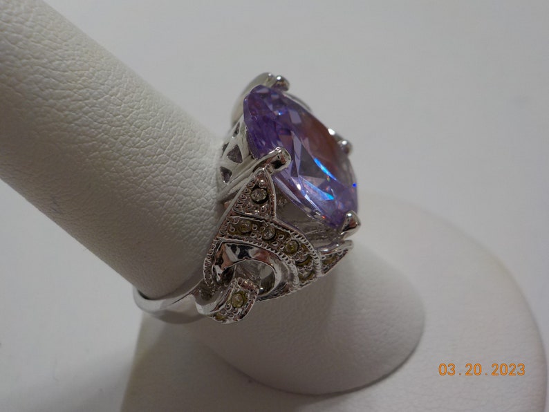 May include: A silver ring with a large, oval-shaped, purple gemstone surrounded by smaller, clear gemstones.