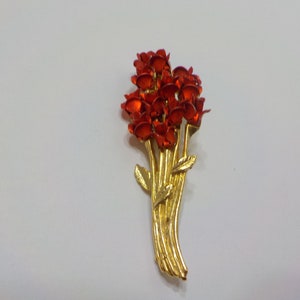 May include: A gold-toned brooch with a cluster of red flower-shaped accents. The brooch is shaped like a bouquet of flowers with stems and leaves.