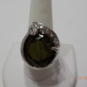 May include: A silver ring with a large, dark green gemstone and smaller clear gemstones. The ring has a unique, curved design.