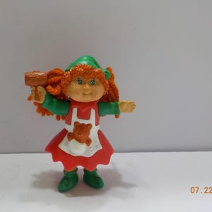 May include: A small plastic doll with orange yarn hair, wearing a green hat, a green long-sleeved shirt, and a red skirt with a white apron featuring a brown bear graphic. The doll holds a brown mallet and has the letters CPK printed on the apron.