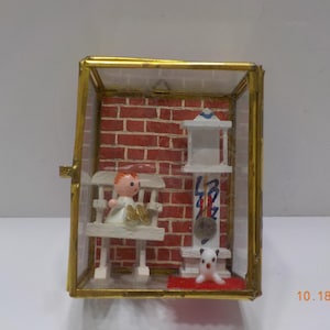 May include: A miniature display case with a gold frame houses a scene with a red brick wall backdrop. Inside, a doll sits on a white bench, and a clock and a dog are also present. The date "10.18.2025" is visible on the bottom right.