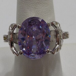 May include: A silver ring with a large, oval-shaped, purple gemstone surrounded by smaller clear gemstones.