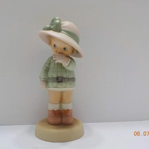 May include: A ceramic figurine of a young child wearing a hat with a green bow, a green plaid jacket, and brown boots. The figurine is standing on a round base. The color palette includes beige, green, and brown.