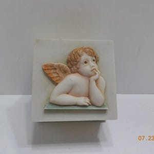 May include: A white ceramic box with a raised image of a cherub with wings. The cherub is looking to the right and has a hand on its chin.