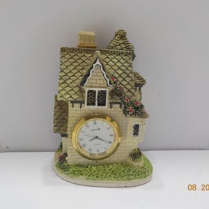 May include: A tabletop clock shaped like a cottage with a beige stone-textured exterior, a green shingled roof, and climbing red flowers. The clock face is circular with a gold-tone rim, white background, black hands, and Roman numerals.