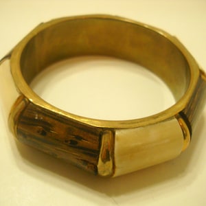 May include: A gold-toned bangle bracelet with a geometric design. The bracelet is made of metal and has a wood-like pattern on the outside. The inside of the bracelet is smooth and polished.