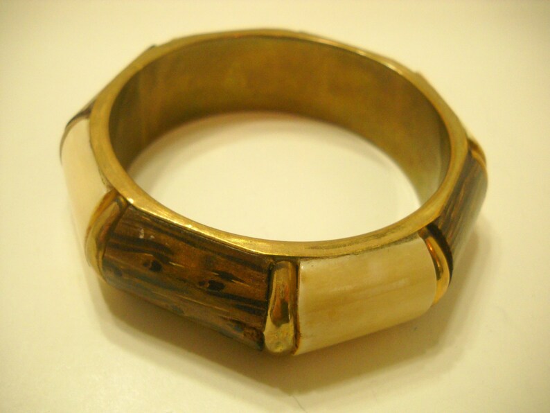 May include: A gold-toned bangle bracelet with a geometric design. The bracelet is made of metal and has a wood-like pattern on the outside. The inside of the bracelet is smooth and polished.