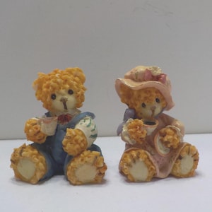May include: Two decorative teddy bear figurines. One bear wears a blue outfit and holds a teacup. The other bear wears a pink dress and a hat, also holding a teacup. Both bears have yellow fur and are seated.