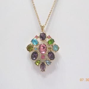 May include: Gold-toned pendant necklace featuring a cluster of colorful gemstones. The pendant has a central pink oval stone surrounded by various colored stones, including purple, blue, green, and pink. The necklace has a delicate gold chain.
