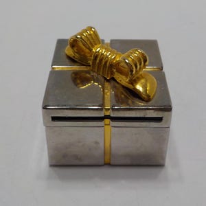 May include: A small silver and gold gift box with a gold bow on top. The box is about 2 inches tall and wide.