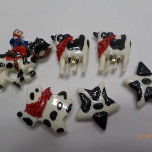 May include: A set of six black and white cow-themed buttons with red accents. The buttons feature a cowboy riding a horse, two cows, a star, and the state of Texas.