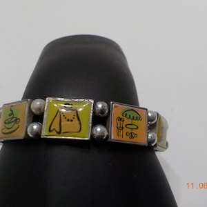 May include: A silver charm bracelet with colorful enamel charms featuring images of a cup of coffee, a dress, a table with an umbrella, and a flower.