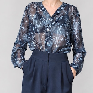 Puede incluir: Una blusa de manga larga con botones en tejido azul marino semitransparente con un estampado floral botánico en azul claro y blanco. La blusa tiene cuello en V y se combina con pantalones azul marino de cintura alta con pliegues frontales.