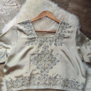 May include: A cream-colored short-sleeve blouse featuring a square neckline and intricate silver embroidery. The garment displays detailed floral and geometric beadwork patterns along the neckline, the center front panel, the hem, and the sleeve cuffs.