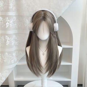 Könnte beinhalten: Ein Schaufensterpuppenkopf mit einer langen, glatten, aschbraunen Perücke mit gestuftem Pony und weißen Over-Ear-Kopfhörern. Die Puppe trägt eine silberfarbene Halskette mit Kreuzanhänger und weist eine neutrale, glatte Gesichtsstruktur auf.