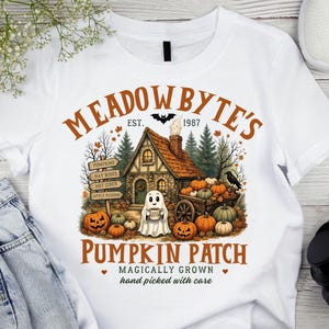 May include: A white t-shirt featuring a graphic of a cottage, a ghost holding a mug, and pumpkins. The text reads MEADOWBYTE'S EST. 1987 PUMPKIN PATCH MAGICALLY GROWN hand picked with care, with signs for PUMPKINS, HAY RIDES, HOT CIDER, and APPLE PICKING.