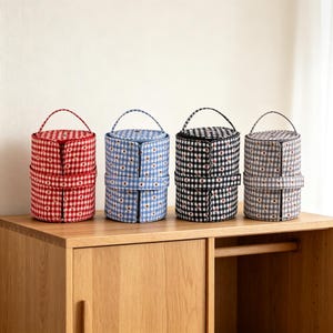 May include: Four cylindrical storage cases with handles, each featuring a gingham pattern with small floral accents. The cases are available in red, blue, black, and gray, and each includes a horizontal strap detail around the center body.