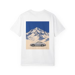 以下が含まれることがあります： 背面に長方形のグラフィックがプリントされた白いTシャツ。グラフィックには、深い青空の下、雪を頂いた山脈を背景に駐車されたシルバーのスポーツカーが描かれています。車のすぐ下には、濃い青色でGROSSGLOCKNERという文字が印刷されています。