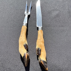 Peut inclure: Un ensemble de découpe composé d'une fourchette et d'un couteau dentelé en métal. Les deux ustensiles possèdent des manches formés par des pattes d'animal couleur fauve, avec des poils sombres et des sabots à la base, fixés par des viroles.