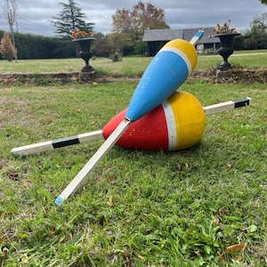 May include: Two large wooden fishing floats with elongated stems. One float features a blue and yellow body, while the other has a red and yellow body. Both stems are painted with black and white sections, and each has a small loop at the end.