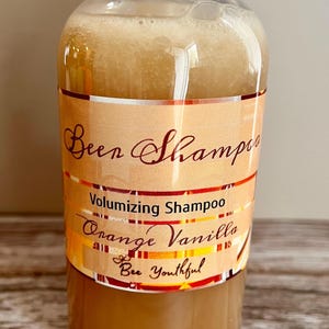 May include: A clear plastic bottle of Bee Shampoo volumizing shampoo in orange vanilla scent. The bottle has a white cap and a label with the text "Bee Shampoo", "Volumizing Shampoo", "Orange Vanilla", and "Bee Youthful".