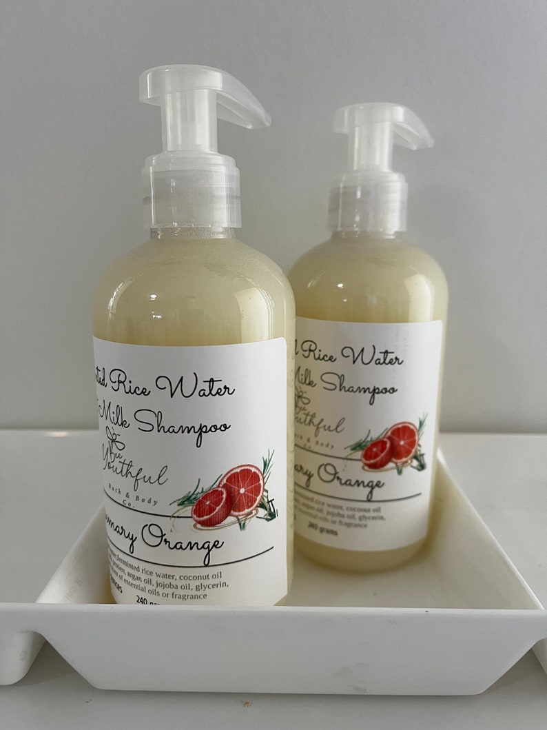 Puede incluir: Dos botellas de champ&uacute; de leche de agua de arroz con aroma a Rosemary Orange, con etiquetas blancas y dispensadores de bomba transparentes. Las etiquetas presentan texto y un gr&aacute;fico de una rodaja de pomelo y ramitas de romero. Cada botella contiene 240 gramos.