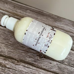 Puede incluir: Una botella de pl&aacute;stico blanco de acondicionador para el cabello con una tapa blanca. La etiqueta dice "Fermented Rice Water, Goat Milk, Nourishing Conditioner, Leave In or Rinse Out, Bee Youthful". La botella est&aacute; sobre una superficie de madera.