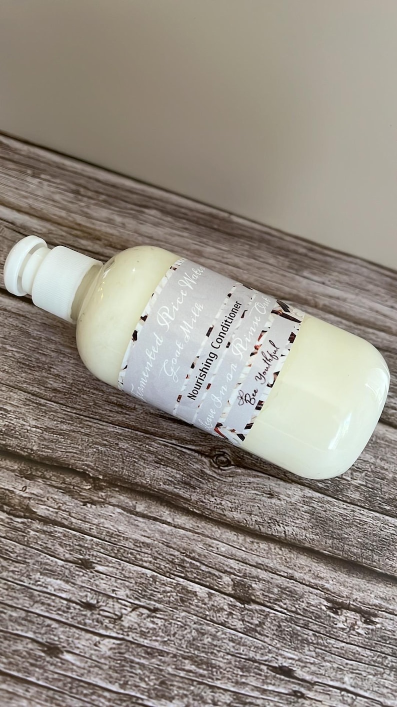 Puede incluir: Una botella de pl&aacute;stico blanco de acondicionador para el cabello con una tapa blanca. La etiqueta dice "Fermented Rice Water, Goat Milk, Nourishing Conditioner, Leave In or Rinse Out, Bee Youthful". La botella est&aacute; sobre una superficie de madera.