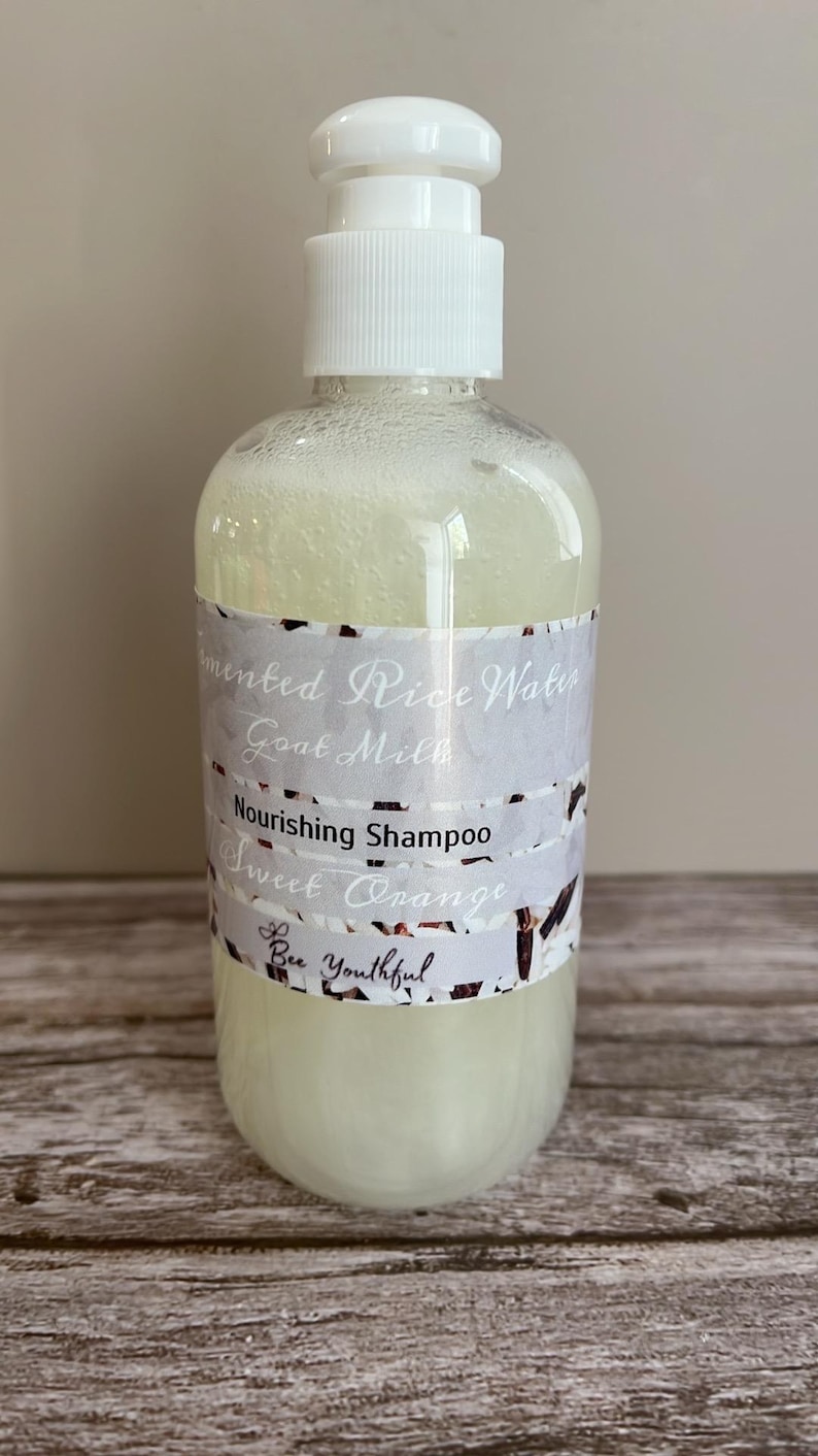 Puede incluir: Una botella de pl&aacute;stico transparente de champ&uacute; con una tapa de bomba blanca. La etiqueta dice "Fermented Rice Water Goat Milk Nourishing Shampoo Sweet Orange" y "Bee Youthful". El champ&uacute; es de color amarillo p&aacute;lido.