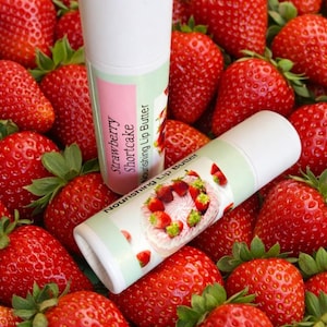 Puede incluir: Dos tubos blancos de manteca labial con etiquetas que dicen "Strawberry Shortcake Nourishing Lip Butter" descansan sobre un lecho de fresas frescas y maduras. Las etiquetas presentan una combinación de colores rosa y verde con un gráfico de pastel de fresas.