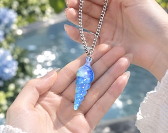 May include: A pendant necklace featuring a spiral-shaped, translucent blue and white glass shell charm suspended from a silver-toned metal curb chain. The charm displays a swirling, marbled colour pattern with a glossy finish and a tapered point.