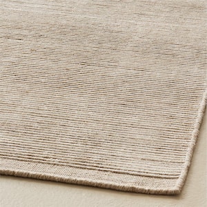 May include: A rectangular area rug featuring a textured, ribbed surface in a neutral beige tone. The rug displays a consistent horizontal linear pattern created by raised fibers, with a finished edge border running along the bottom side.