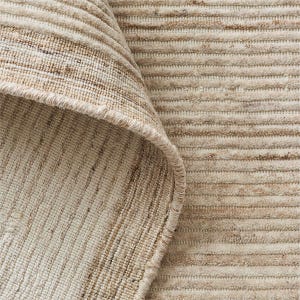 May include: A textured area rug featuring a repeating pattern of horizontal raised ridges in shades of beige, cream, and light brown. The rug is folded over at the top left corner, revealing the woven underside and the finished edge binding.
