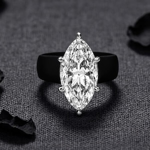 May include: A ring featuring a large, clear, marquise-cut gemstone set in a silver-toned metal prong setting, mounted on a smooth, solid black band. The faceted stone reflects light, creating a bright, sparkling appearance against the dark band.