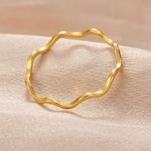 Può includere: Un anello sottile color oro con un design ondulato. Il metallo ha una finitura opaca e spazzolata, che crea una consistenza morbida lungo la fascia circolare. L'anello presenta un motivo curvo continuo e ripetitivo che forma una sagoma.