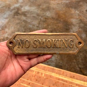 Puede incluir: Un letrero rectangular de latón con un acabado dorado oscuro texturizado que presenta el texto en relieve NO SMOKING en letras mayúsculas. El letrero tiene una forma hexagonal puntiaguda en ambos extremos con un orificio de montaje circular.