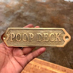 Könnte beinhalten: Ein rechteckiges Messingschild mit strukturiertem Hintergrund und glatter, erhabener Schrift mit der Aufschrift POOP DECK. Das Schild hat eine spitz zulaufende, hexagonale Form mit zwei vorgebohrten Löchern an den Seiten zur Befestigung.