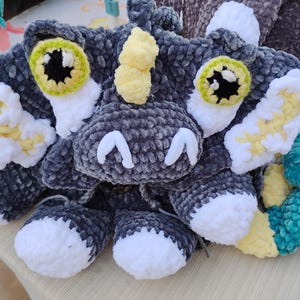 May include: A plush crochet creature made of dark gray, white, and pale yellow yarn. The creature features large, round eyes with lime green irises, a small yellow horn on its head, and white accents on its ears, feet, and snout area.