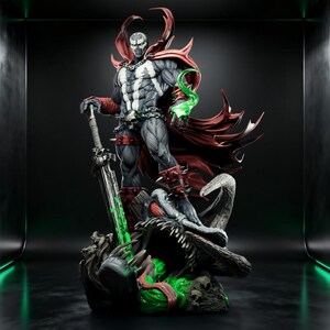 Op de afbeelding: Een gedetailleerd Spawn-figuur met een gespierd lichaam in een zwart-wit pak met een wapperende rode cape, zilveren kettingen en armbanden met stekels. Het figuur houdt een groot zwaard vast en staat op een voetstuk met groene energie-effecten.
